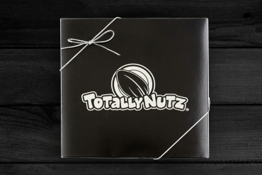 black square box with silver totally nutz logo embossed on top and closed with a silver ribbon, box has gingham print bottom and the photo is taken on a white wood background 